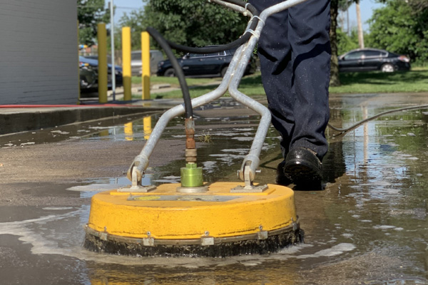 Power washing technician using circular surface cleaner to wash concrete behind a business building