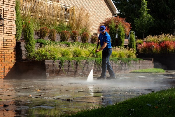 Power washing technician washing a stone patio with foliage and a brick house in the background