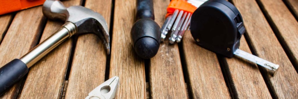 Just a few of the 21 tools every homeowner should have, including as a hammer, screwdriver, and measuring tape, set on the surface of a wooden deck