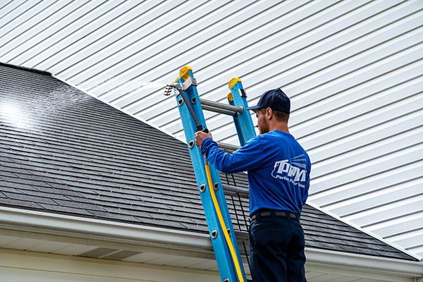 Professional power washing technician performing a roof treatment service from a ladder.
